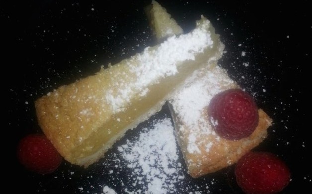 Two triangular slices of a pale cake dusted with powdered sugar and garnished with raspberries on a dark plate