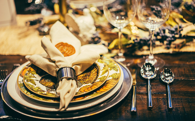 Close-up of an elegant table setting with stacked decorative plates, a folded cream napkin in a ring, and wine glasses in warm gold and cream tones