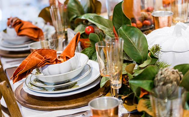 Table setting with white bowls and plates, an orange folded napkin, greenery and small red tomatoes as decoration on a wooden surface