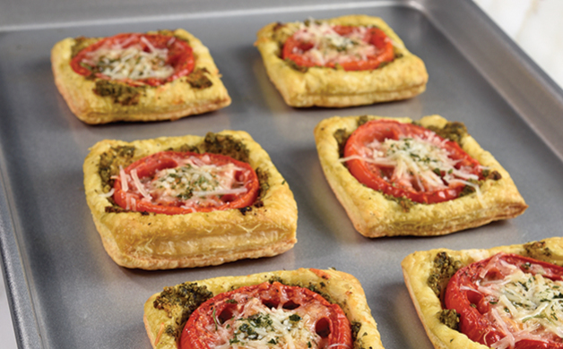 Six square puff pastry tarts topped with tomato slices, pesto, and grated cheese on a metal baking tray, warm reddish tones