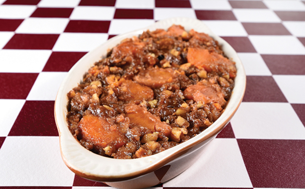 A dish of candied sweet potatoes with nuts and brown sugar glaze in a white oval baking dish, set on a red and white checked cloth