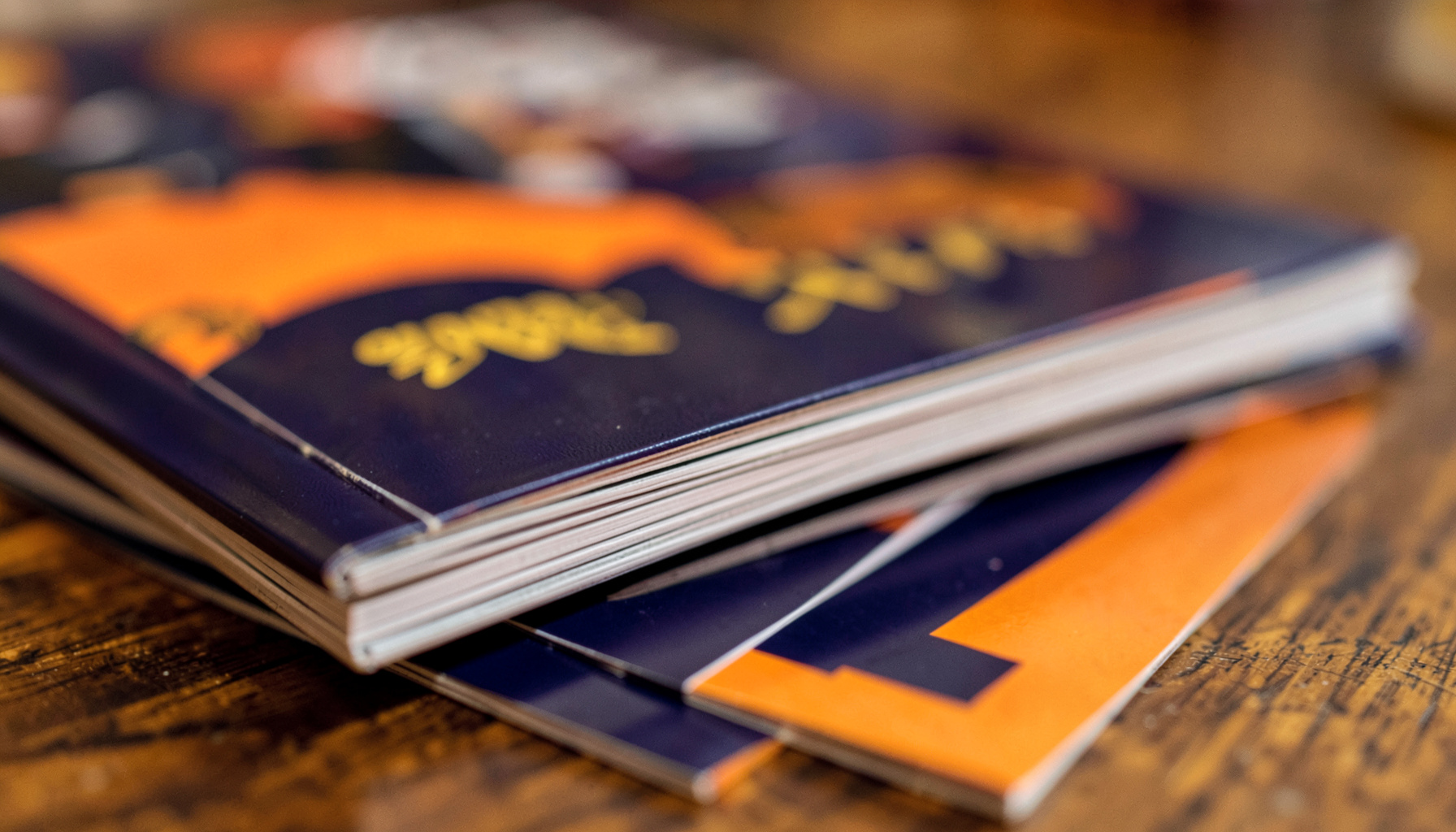Soft-focus photograph of glossy magazine covers stacked on a wooden table, rich navy and gold tones