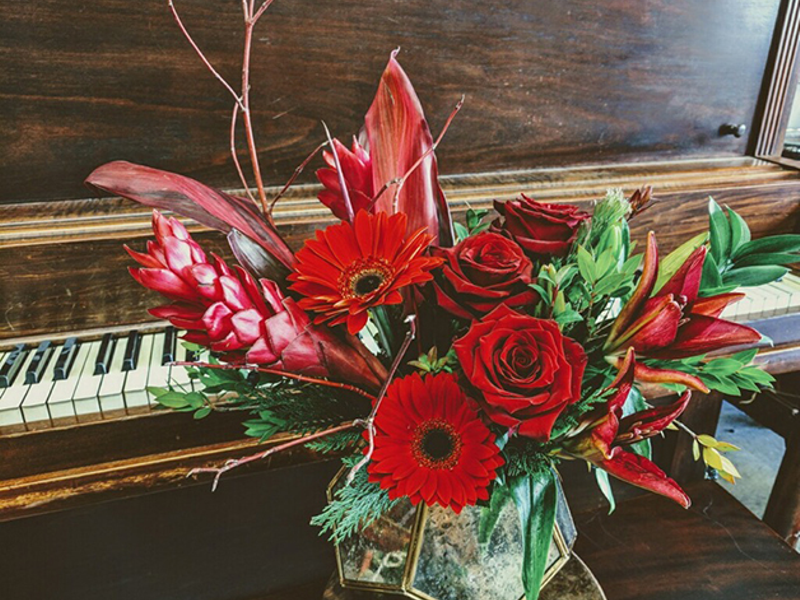 A red and green floral arrangement with roses, gerbera daisies, and tropical leaves resting on an upright piano keyboard, warm and celebratory mood